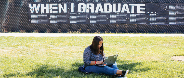 A woman sitting on the grass outside, working on her laptop. Behind her is a sign that reads, "WHEN I GRADUATE."