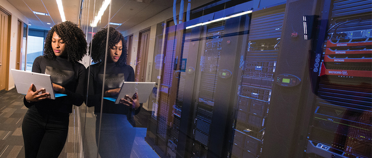 A woman holding a laptop standing in a hallway leans against a wall of mirrors, behind which are several blocks of IT network servers and equipment.